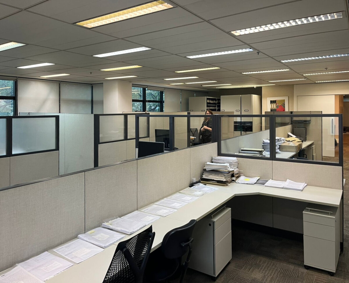 Foto interna de escritório mobiliado, com divisórias a meia altura, luminárias embutidas no forro e com amplas janelas.